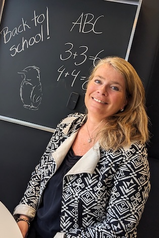 Hvit kvinne med en svart tavle i bakgrunnen med skriften Back to school skrevet med hvitt kritt.