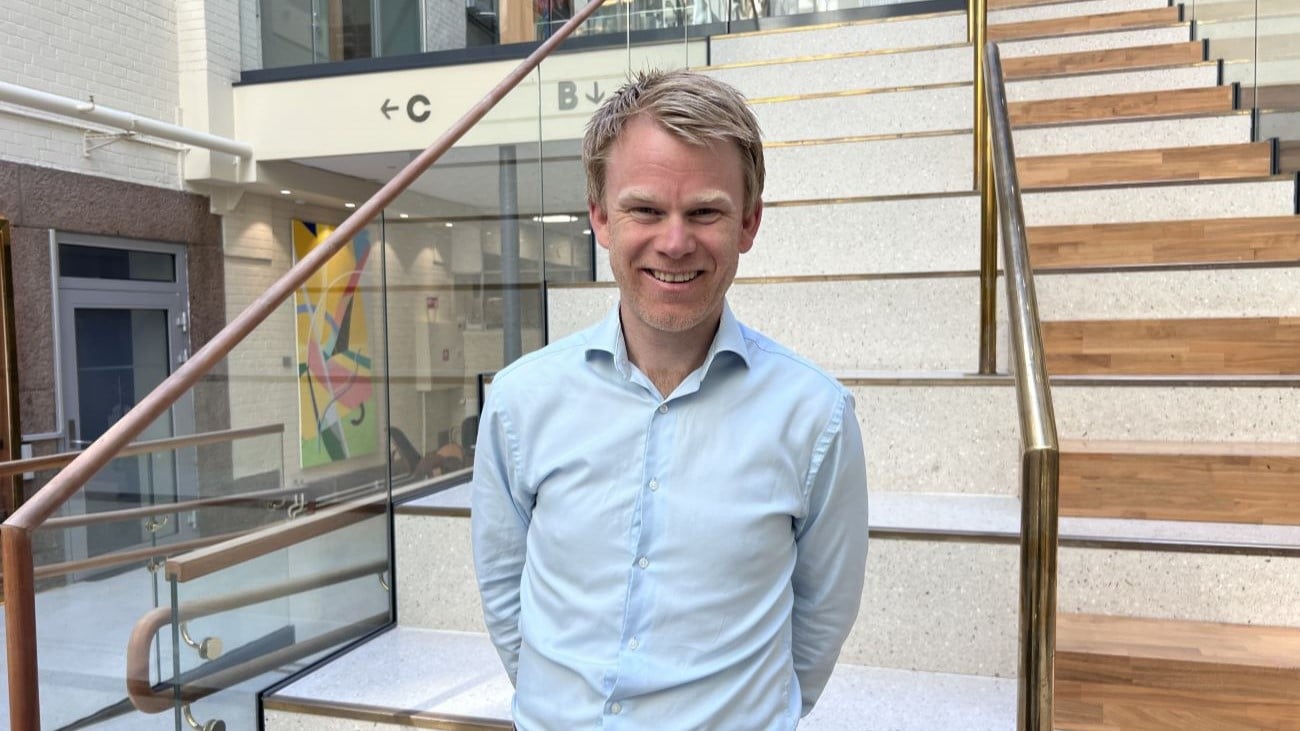 Mann som står foran en trapp i et kontorlokale og smiler bredt til kamera.