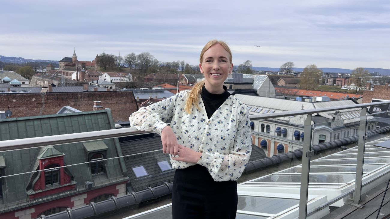 Ung kvinne som står på en takterrasse i Oslo og smiler bredt til kamera.