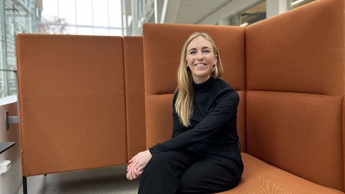 Ung kvinne som sitter i en stor oransje sofa i et kontorlandskap og smiler til kamera.