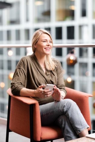 Kvinne som sitter i en loungestol i et lyst lokale mens hun holder mobilen sin og smiler.