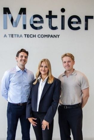 To menn og en kvinne ansatt i Metier som står sammen foran en hvit vegg med teksten "Metier" på.