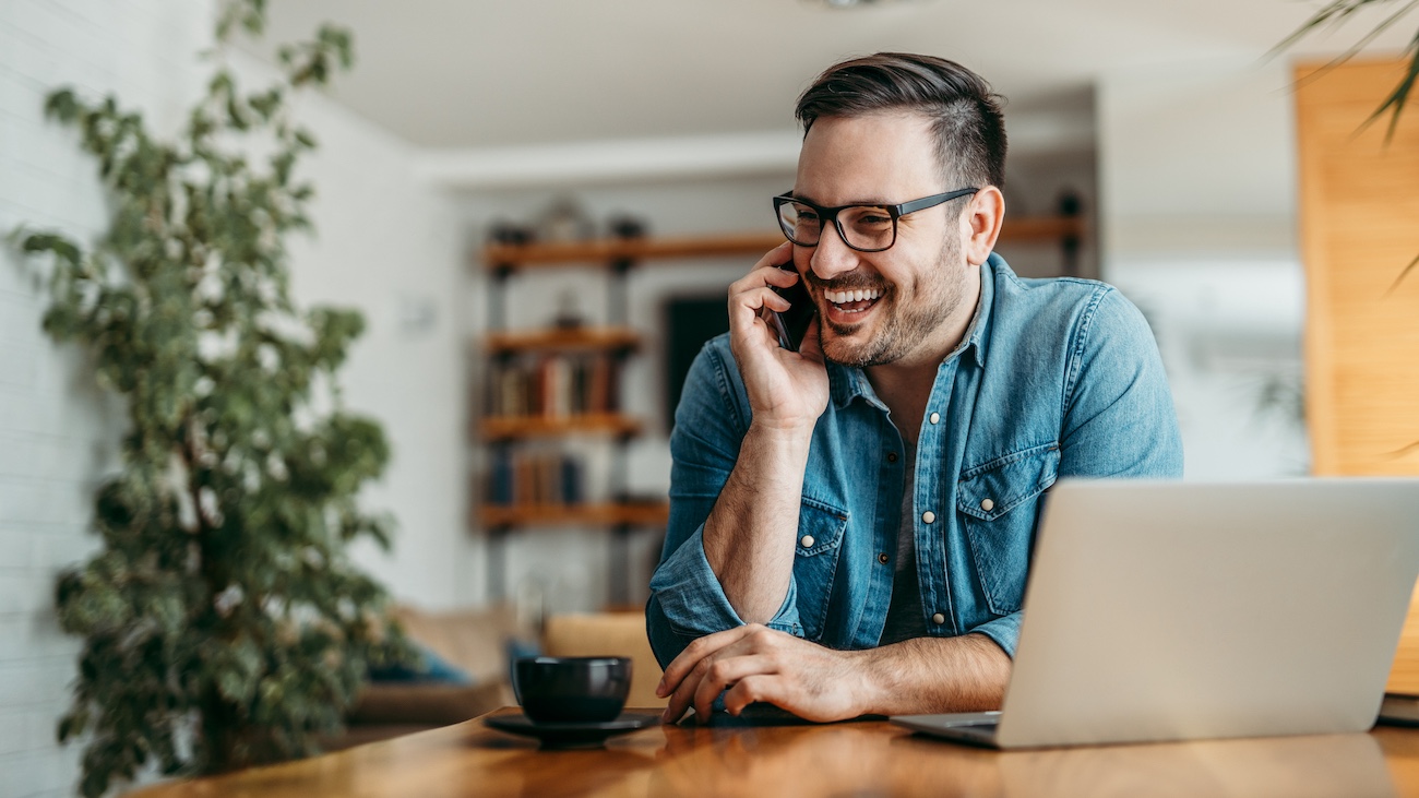 En smilende mann snakker i telefonen mens han sitter ved et bord med en bærbar PC og en kopp kaffe, i et lyst og moderne kontormiljø. Bildet formidler en følelse av karriererådgivning og glede over å utforske muligheter for en ny jobb, knyttet til Econa.