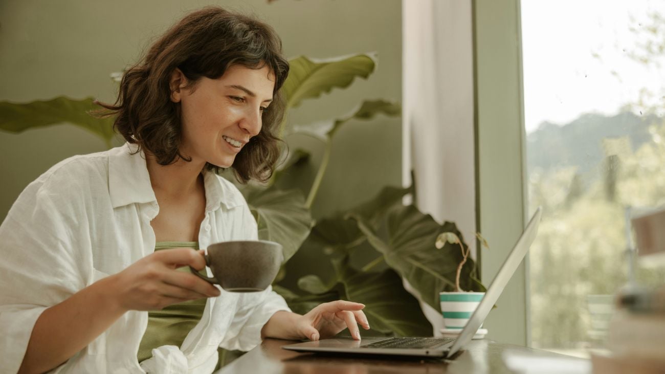 Ung kvinne som sitter og holder i en kaffekopp mens hun ser på PC-skjermen sin og smiler.