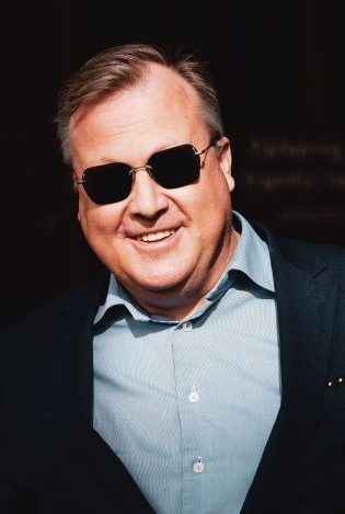 Portrettfoto av smilende mann i dress og mørke solbriller.