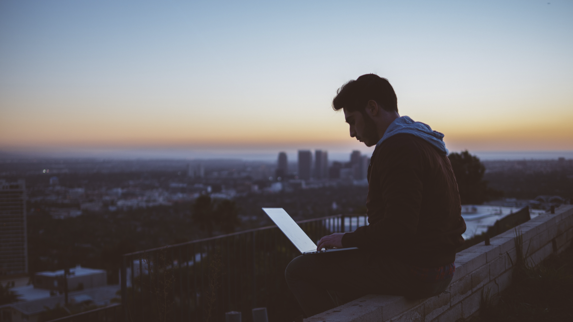Ung mann som sitter på toppen av en bygning med laptopen i fanget og solnedgang i det fjerne.