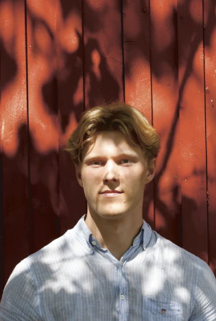 Portrettfoto av en ung mann som står foran en rød husvegg og ser inn i kamera med et seriøst uttrykk.