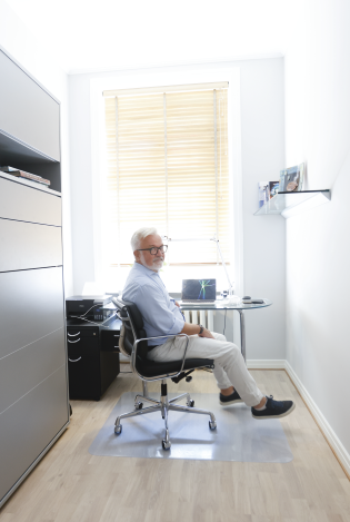 Eldre mann som sitter på en kontorstol i et lyst kontor, snudd vekk fra laptopen og smiler inn i kamera.