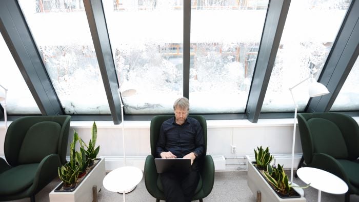 Mann som sitter med en laptop i fanget sitt i et lyst kontorlandskap med store vinduer. 