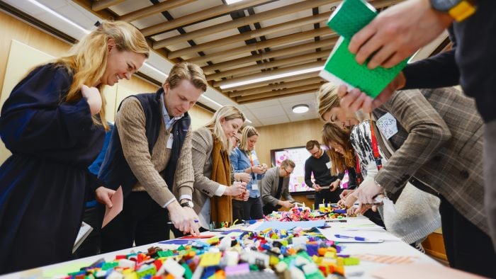 En gruppe mennesker som står rundt et langbord og løser en oppgave ved hjelp av fargerike legoklosser.