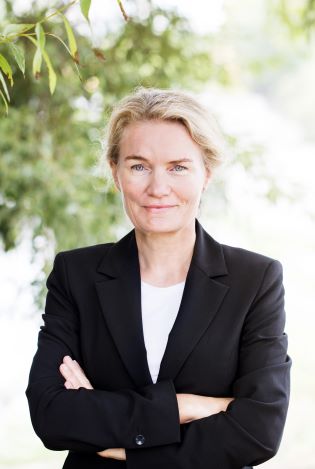 Formelt kledd kvinne som står med armene i kryss foran et tre og ser bestemt inn i kamera.