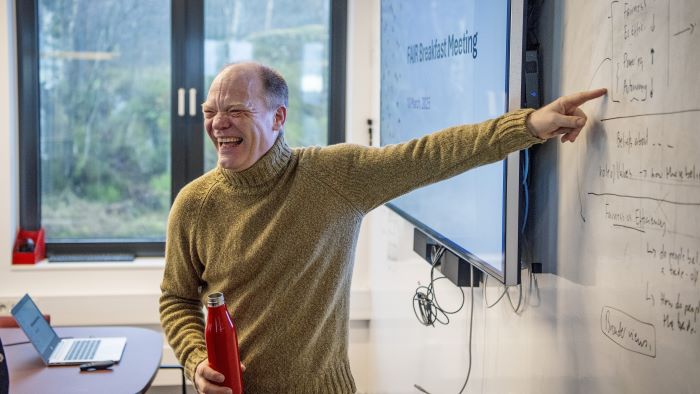 Mann som står i et klasserom og peker på tekst på tavlen mens han ler høyt.