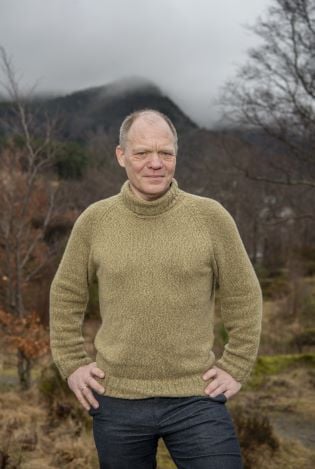 Mann som står i skogen og hviler hendene på hoftene mens han smiler forsiktig til kamera.